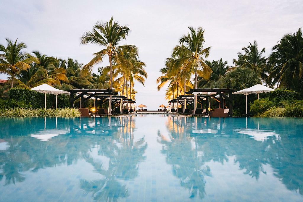 Teaser Bild Blick über das Wasser eines Pools umgeben von Palmen bei Sonnenuntergang